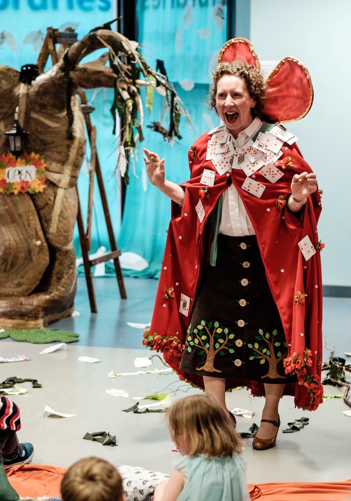 An image from the production Alice and the Library Tree. In the background on the left side you can see a tree, made from fabric, looking old and gnarled. A sign hung on the tree, with a border made from Autumn leaves, says OPEN. There is an old fashioned lamp above the sign, and what looks like a ladder just behind the tree. Green and white ribbons hang from the tree's branches.

In front of the tree, to the right, is a singer, mid-song who wears a red cape decorated with playing cards, all from the heart suit and roses.. Her black skit is decorated with appliqued trees. She also wears what appear to be red and gold wings behind her head.

There are scraps of material, debris from the tree on the floor around her, and to the foreground of the picture you can see the heads of the young audience.