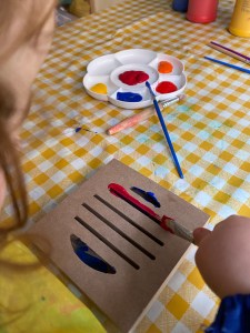 The image shows a child painting a wooden block instrument. The instrument, being painted with red and blue is in the foreground with a set of paints on a small white palette in the background. 