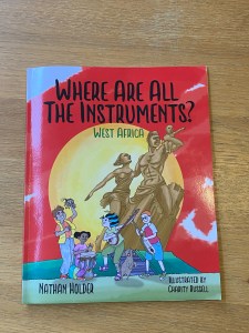 The front cover of the book that is the subject of this review. It is a red book and you can see the children from the Why Squad enjoying playing musical instruments from West Africa in front of a statue of the African Renaissance Monument.
