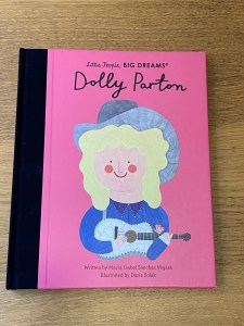 The book’s front cover, shown here, has a cartoon image of Dolly Parton on a bright pink background. Dolly Partong wears a grey cowboy hat and carries a guitar. 
