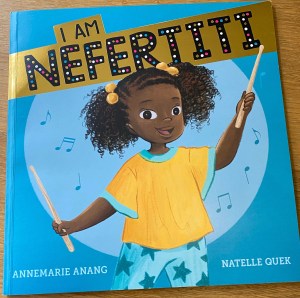 Showing the front cover of the book. A little Black girl, Nefertiti, happily and proudly holds her drumsticks ready to play.