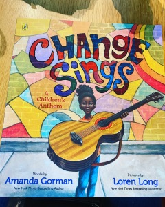 The front cover of the book Change Sings. This is a beautiful, brilliantly vibrant and colourful front cover with the title Change Sings, subheading A Children's Anthem, and the author and illustrators' names noted. The main illustration is a young, black girl stood holding a large guitar out in front of herself. She stands on a sidewalk with a colourful mural behind her.