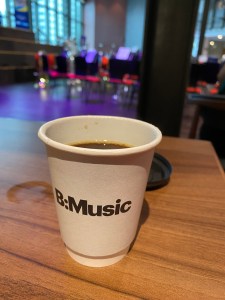 A coffee cup showing the B:Music logo sits on a table. In the background you can see the stage area set with chairs and music stands ready for the performance.