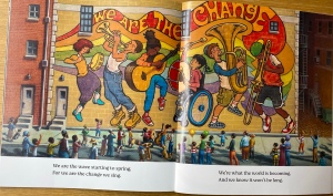 A beautiful, bright, joyful, multi-coloured picture showing a billboard style poster of the children all playing their instruments together.  A banner is behind the children in this poster saying “We are the change”. There is a crowd of people looking at the poster. 