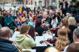 The viewer is seated within the orchestra looking towards the conductor and the crowd watching them perform. The strings have their bows raised,  and the brass players have their instruments to their lips ready to play. 