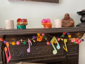 Brightly coloured bunting made out of musical notation hung at our black fireplace. There are some objects on the mantlepiece above including two brightly coloured elephant candle holders and a candle in the shape of a buddha. The musical bunting is accompanied by a pom pom garland.