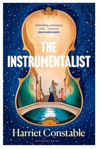 This picture shows the front cover of the book The Instrumentalist by Harriet Constable. The cover has a dark blue, night sky background complete with stars. On top of this background is the form of a violin which also has a picture of a girl playing a violin while standing on a simple bridge in Venice. There are tall buildings either side of the bridge with a canal flowing under the bridge.
