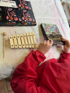 My little girl, dressed in an art apron, opening the packet of metallic acrylic paint pens with her plain wooden xylophone and beaters in front of her ready to decorate. I have put out a table cloth that we always use for any art projects on our kitchen table to protect it.