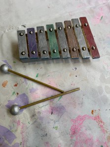 My daughter's finished and decorated xylophone. She chose to paint each of the tone bars a different metallic colour, and the head of the beater in silver metallic paint. The xylophone sits on our very well used art table cloth and so has a lot of paint stains all over it.