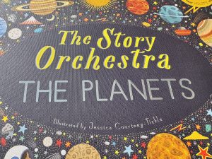 The cover of the book The Story Orchestra: The Planets. It has a black cover, fitting for a book about space, and illustrations of planets, stars, moons, alien ships and even what looks like a snow topped mountain on.